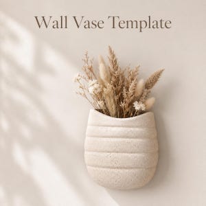 May include: A beige wall vase template with a textured, ribbed surface holding a bouquet of dried grasses and small white flowers. The text Wall Vase Template is printed in dark brown above the vase, which is mounted on a neutral wall.