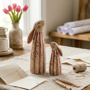 May include: Two ceramic rabbit figurines with red and white striped bodies and long ears. The figurines are decorated with floral accents. A vase of pink tulips and a set of mugs are in the background.