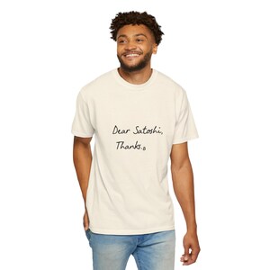 Könnte beinhalten: Cremefarbenes T-Shirt mit dem handgeschriebenen Text "Dear Satoshi, Thanks." Das Shirt ist ein lässiger Stil mit kurzen Ärmeln. Die Person auf dem Bild trägt eine blaue Jeans.