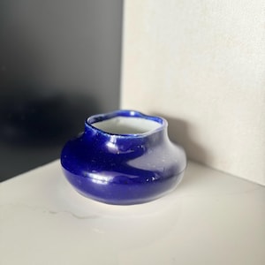 May include: A small, round, cobalt blue ceramic vase with a wide body and a narrow, scalloped rim. The interior is white. The vase has a glossy finish and sits on a white surface. The background is a mix of white and dark gray.