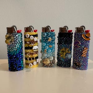 May include: Five decorative lighters with jewel and bead embellishments. The lighters feature a variety of colors, including blue, yellow, and purple, and are adorned with charms and decorative elements.