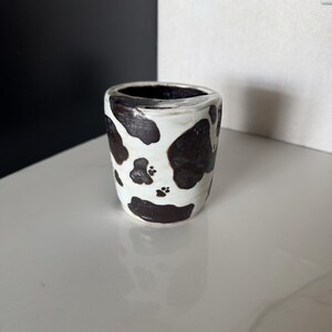Peut inclure: Une petite tasse cylindrique en céramique avec une base blanche et des taches noires irrégulières rappelant un motif de vache. La surface présente un aspect texturé et artisanal avec un intérieur sombre et des détails de pattes.