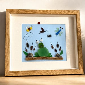 Peut inclure: Une œuvre d'art murale encadrée représentant une grenouille en verre de mer vert assise sur un morceau de bois flotté. La scène comprend des insectes en verre de mer, une petite coccinelle rouge et des massettes brunes en pierre et fil de fer.
