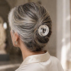 May include: A silver-toned hair clip in a spiral design, set against gray and white hair. The clip is the focal point, adding a touch of elegance. The person is wearing a cream-colored satin blouse.
