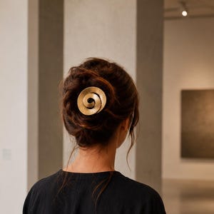 Op de afbeelding: Een goudkleurig haaraccessoire met een cirkelvormig, spiraalvormig ontwerp. Het metalen stuk is bevestigd in een donkerbruin opgestoken kapsel. Het accessoire heeft een getextureerde, matte afwerking en is centraal achter op het hoofd geplaatst.