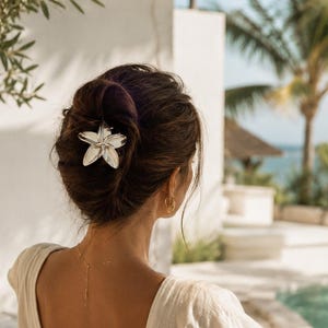 Op de afbeelding: Een vrouw met donker haar in een knot, versierd met een zilveren bloemvormige haarclip. Ze draagt een crèmekleurige jurk met een lage rug. De scène speelt zich buiten af, met uitzicht op een zwembad en palmbomen.