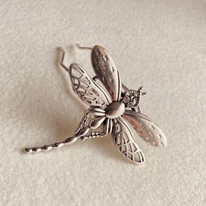 May include: A silver-toned dragonfly brooch with intricate wing detailing and a textured body. The brooch features a pin clasp on the back for secure attachment. The dragonfly's wings have a delicate, openwork design.