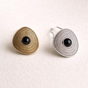 Puede incluir: Dos pendientes, uno dorado y triangular, el otro plateado y circular. Ambos presentan un diseño en espiral y una piedra central negra y circular. Los pendientes se muestran sobre una superficie blanca.