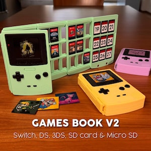 May include: A light green folding game storage case shaped like a handheld console, containing multiple game cartridges and numbered slots. Includes a yellow console-shaped case, a pink one, and small character stickers. Text reads GAMES BOOK V2.