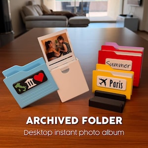 May include: A set of desktop instant photo albums shaped like miniature file folders in light blue, white, yellow, red, and pink. The folders feature labels with text like Summer and Paris, and icons including a heart and an airplane.