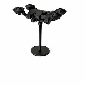 May include: A black model of a spacecraft, featuring four circular engine components, is mounted on a black stand. The spacecraft has a detailed design with various geometric shapes and a small red accent. The stand has a circular base.