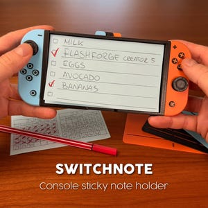 Puede incluir: Un soporte de notas adhesivas de plástico negro con forma de consola de juegos portátil, con un mando izquierdo azul claro y un mando derecho naranja. El centro sostiene una lista de papel rectangular con texto escrito a mano: MILK, FLASHFORGE CREATOR 5, EGGS, AVOCADO, BANANAS.