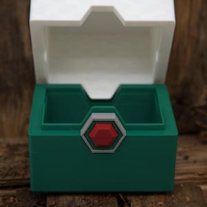 Op de afbeelding: Een groene doos met een wit deksel, open om de binnenkant te onthullen. De doos heeft een rood en grijs embleem op de voorkant. De doos is gemaakt van plastic en staat op een houten oppervlak.