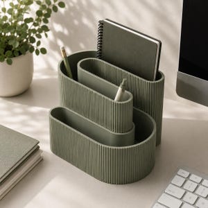 Può includere: Un organizer da scrivania color verde salvia caratterizzato da un design ovale a più livelli con una texture a coste verticali. L'organizer presenta tre scomparti distinti di altezze variabili, contenenti un taccuino a spirale e due penne.