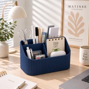 Puede incluir: Un organizador de escritorio azul marino con textura acanalada vertical, que cuenta con múltiples compartimentos llenos de artículos de papelería, incluyendo tijeras, bolígrafos, un cuaderno de espiral con la etiqueta SODA CAT y un estuche blanco.