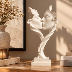 Può includere: Una scultura bianca che raffigura due volti di profilo, posizionati come se si stessero baciando. I volti sono incorniciati da bordi fluidi a forma di petalo che si fondono in una base unica. La scultura è montata su un piedistallo quadrato a gradini.