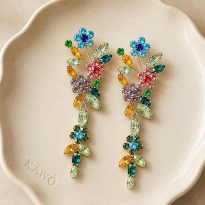 Peut inclure: Une paire de longues boucles d'oreilles pendantes au motif floral, composées de cristaux multicolores dans des tons bleu, vert, jaune, rose et violet. Les boucles d'oreilles présentent une structure verticale en forme de branche avec des feuilles.