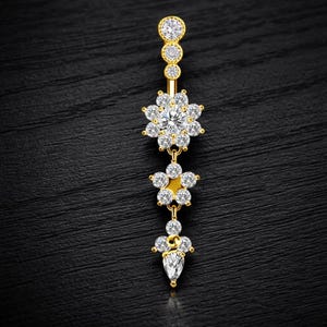 Pode incluir: Um piercing de umbigo dourado com um design floral. A parte superior tem três pequenas pedras preciosas redondas e transparentes. O centro apresenta uma flor com pedras preciosas transparentes. A parte inferior tem uma flor menor e uma gema em forma de gota.