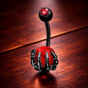 Puede incluir: Un piercing para el ombligo negro con una gema roja en la parte superior y un orbe rojo y negro sostenido por una mano de esqueleto negro. La joya está sobre un fondo de vetas de madera oscura.