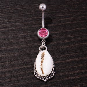 Puede incluir: Un piercing para el ombligo plateado con una gema rosa y un colgante de concha blanca. La concha tiene una superficie texturizada y detallada y está enmarcada por un borde plateado decorativo. El anillo tiene un diseño de barra curva.