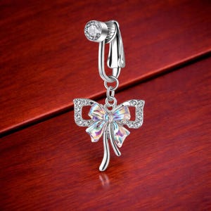May include: A silver-colored belly button ring with a bow-shaped charm. The charm features a shimmering, iridescent center and is outlined with clear, sparkling crystals. The top of the ring has a round, clear crystal.