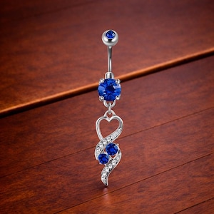 Op de afbeelding: Een zilveren navelpiercing met een gebogen barbell en een bungelend ontwerp. De ring heeft een grote, ronde, blauwe edelsteen aan de bovenkant en twee kleinere blauwe edelstenen met heldere kristallen. Een hartvorm is verwerkt.