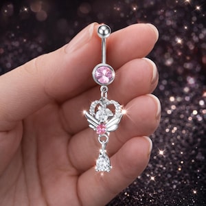 Puede incluir: Un piercing de ombligo plateado con una gema rosa y un dije colgante. El dije presenta una corona, alas y una gema transparente en forma de lágrima. El anillo se sostiene en una mano sobre un fondo oscuro y brillante.