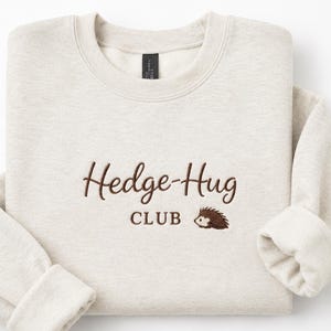 May include: A sand-colored crewneck sweatshirt featuring dark brown embroidered text that reads Hedge-Hug CLUB, accompanied by a small, detailed embroidery of a brown hedgehog positioned to the right of the word CLUB.