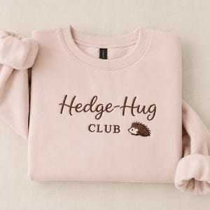 May include: A light pink crewneck sweatshirt featuring dark brown embroidery on the chest. The text reads Hedge-Hug CLUB in a cursive and block font combination, accompanied by a small embroidered graphic of a hedgehog facing to the left.