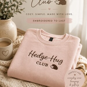 May include: A light pink crewneck sweatshirt featuring dark brown embroidery on the chest that reads Hedge-Hug CLUB with a small hedgehog graphic. The garment has a ribbed collar and cuffs, and the fabric appears to have a soft, fleece-lined interior.