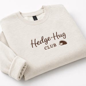 May include: A light beige crewneck sweatshirt featuring dark brown embroidered text that reads Hedge-Hug CLUB, accompanied by a small embroidered graphic of a hedgehog positioned to the right of the word Hug on the front of the garment.