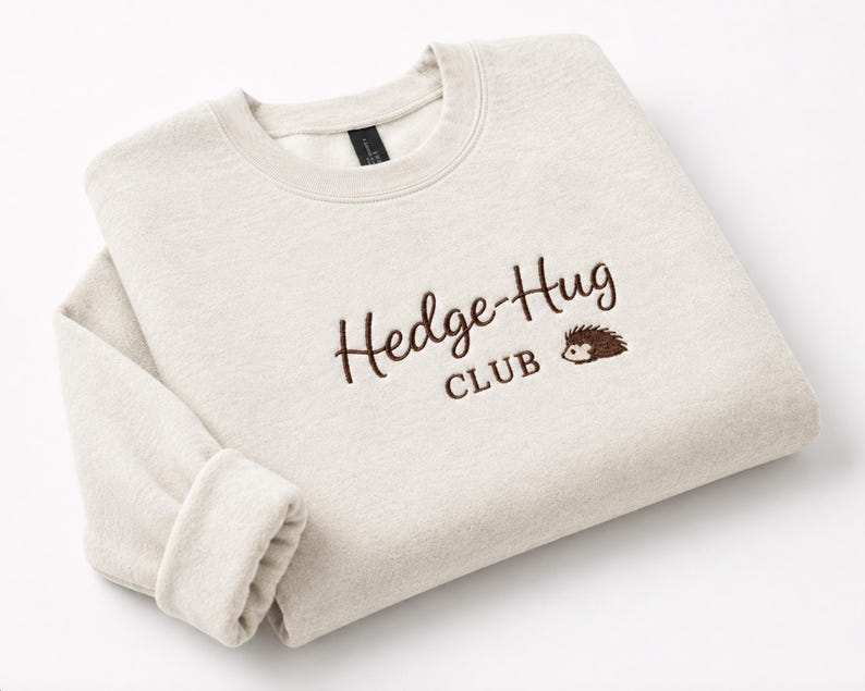 May include: A light beige crewneck sweatshirt featuring dark brown embroidered text that reads Hedge-Hug CLUB, accompanied by a small embroidered graphic of a hedgehog positioned to the right of the word Hug on the front of the garment.