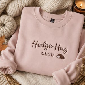 May include: A light pink crewneck sweatshirt featuring dark brown embroidery on the chest that reads Hedge-Hug CLUB with a small embroidered hedgehog icon. The garment has long sleeves and a ribbed neckline, presented in a folded, flat layout.