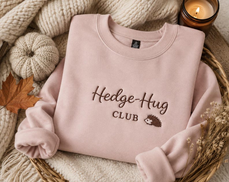 May include: A light pink crewneck sweatshirt featuring dark brown embroidery on the chest that reads Hedge-Hug CLUB with a small embroidered hedgehog icon. The garment has long sleeves and a ribbed neckline, presented in a folded, flat layout.