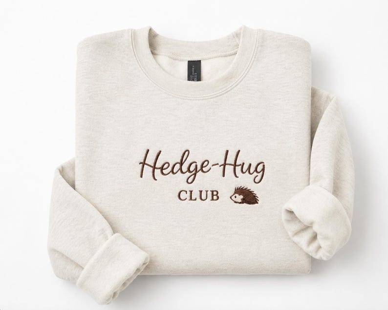 May include: A sand-colored crewneck sweatshirt featuring dark brown embroidered text that reads Hedge-Hug CLUB, accompanied by a small, detailed embroidery of a brown hedgehog positioned to the right of the word CLUB.
