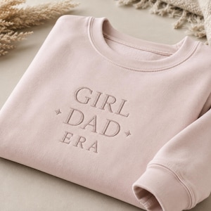 Può includere: Una felpa girocollo rosa chiaro con il testo ricamato GIRL DAD ERA in filo rosa abbinato. Il design include piccoli accenti a forma di stella a quattro punte su entrambi i lati della parola DAD, centrati sul petto del capo.