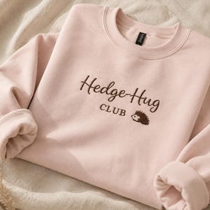 May include: A light pink crewneck sweatshirt featuring dark brown embroidery on the chest. The text reads Hedge-Hug CLUB, accompanied by a small embroidered graphic of a hedgehog. The garment has long sleeves and a ribbed collar neckline.
