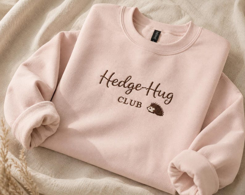 May include: A light pink crewneck sweatshirt featuring dark brown embroidery on the chest. The text reads Hedge-Hug CLUB, accompanied by a small embroidered graphic of a hedgehog. The garment has long sleeves and a ribbed collar neckline.