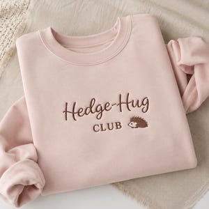 May include: A light pink crewneck sweatshirt featuring dark brown embroidery on the chest. The text reads Hedge-Hug CLUB in a combination of cursive and block lettering, accompanied by a small embroidered graphic of a brown hedgehog.