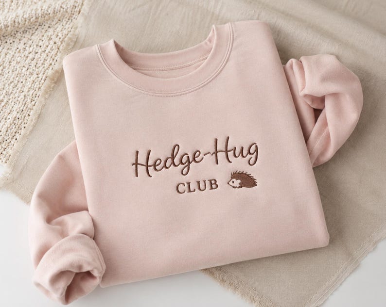 May include: A light pink crewneck sweatshirt featuring dark brown embroidery on the chest. The text reads Hedge-Hug CLUB in a combination of cursive and block lettering, accompanied by a small embroidered graphic of a brown hedgehog.