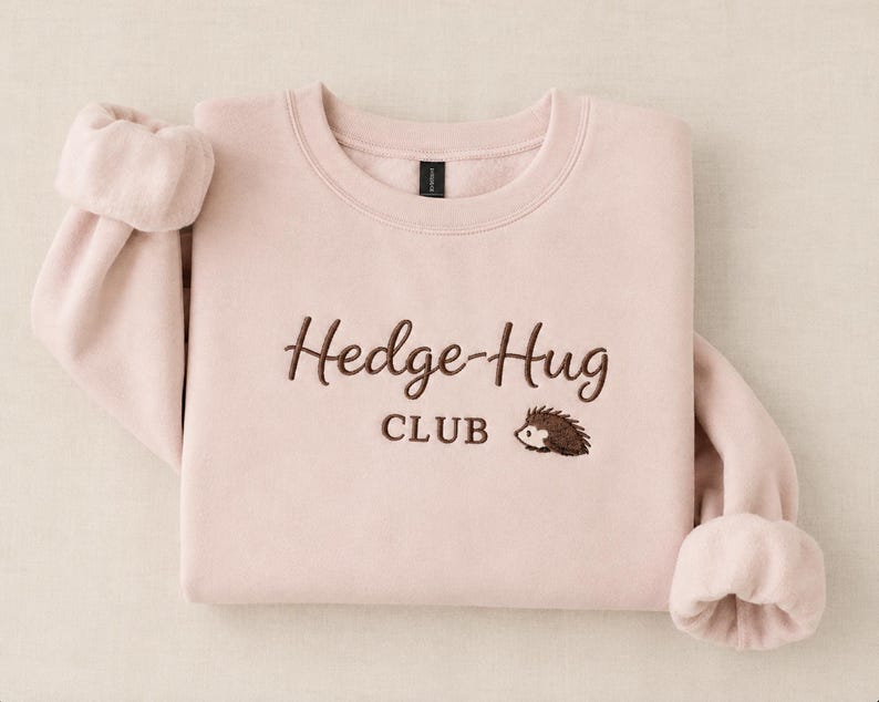 May include: A light pink crewneck sweatshirt featuring dark brown embroidery on the chest. The text reads Hedge-Hug CLUB in a cursive and block font combination, accompanied by a small embroidered graphic of a hedgehog facing to the left.