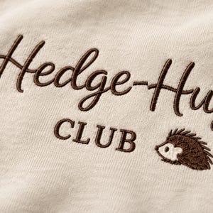 May include: A light beige fabric featuring dark brown embroidery. The text reads Hedge-Hug CLUB in a combination of cursive and block lettering. A small, embroidered brown and white hedgehog icon is positioned to the right of the word CLUB.