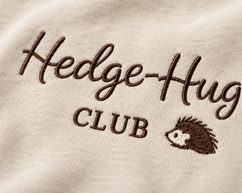 May include: A light beige fabric featuring dark brown embroidery. The text reads Hedge-Hug CLUB in a combination of cursive and block lettering. A small, embroidered brown and white hedgehog icon is positioned to the right of the word CLUB.