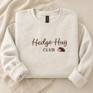 May include: A light heather beige crewneck sweatshirt featuring dark brown embroidery on the chest. The text reads "Hedge-Hug CLUB" in a mix of script and block lettering, accompanied by a small embroidered graphic of a brown hedgehog.