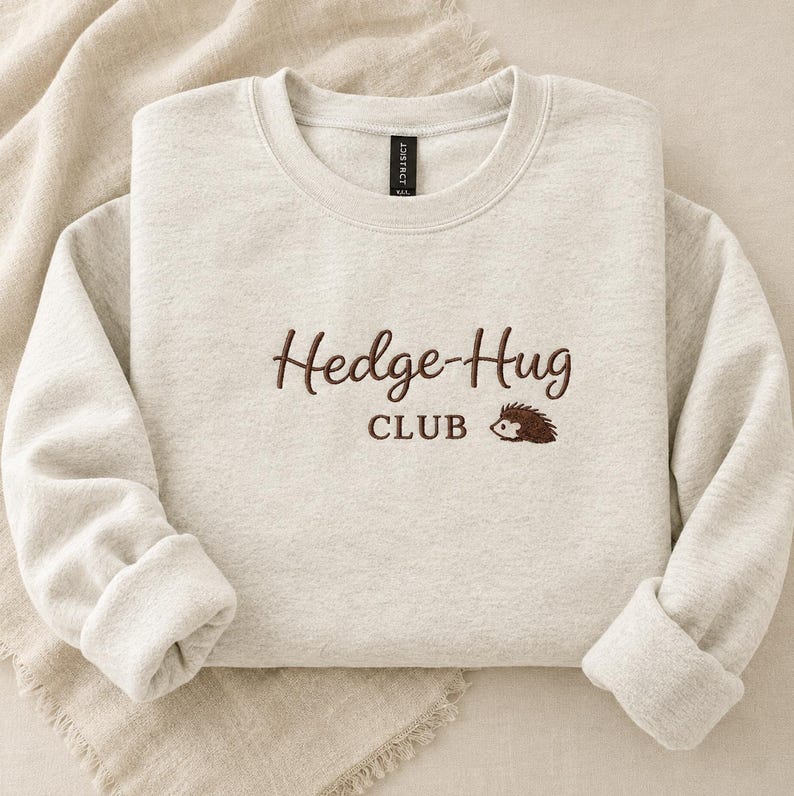 May include: A light heather beige crewneck sweatshirt featuring dark brown embroidery on the chest. The text reads "Hedge-Hug CLUB" in a mix of script and block lettering, accompanied by a small embroidered graphic of a brown hedgehog.