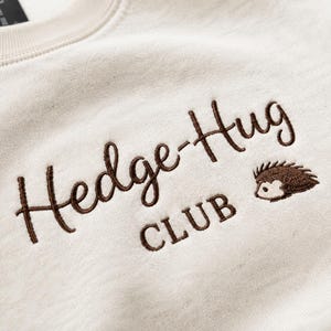 May include: A cream-colored sweatshirt featuring dark brown embroidered text that reads Hedge-Hug CLUB. To the right of the text is a small, detailed embroidery of a brown hedgehog with a light-colored face and dark brown quills.