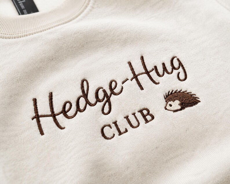 May include: A cream-colored sweatshirt featuring dark brown embroidered text that reads Hedge-Hug CLUB. To the right of the text is a small, detailed embroidery of a brown hedgehog with a light-colored face and dark brown quills.