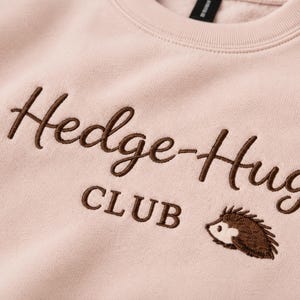 May include: A light pink sweatshirt featuring dark brown embroidered text that reads Hedge-Hug CLUB. Below the text is a small, detailed embroidery of a brown hedgehog facing left. The fabric has a soft, fleece-like texture visible throughout.