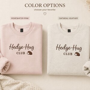May include: Two crewneck sweatshirts in rosewater pink and oatmeal heather colors. Each features dark brown embroidered text that reads Hedge-Hug CLUB with a small brown hedgehog graphic. The sweatshirts have long sleeves and ribbed necklines.