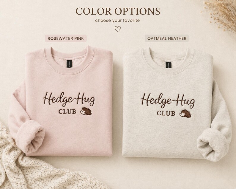 May include: Two crewneck sweatshirts in rosewater pink and oatmeal heather colors. Each features dark brown embroidered text that reads Hedge-Hug CLUB with a small brown hedgehog graphic. The sweatshirts have long sleeves and ribbed necklines.